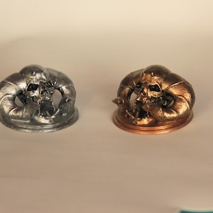 May include: Two small, detailed skull sculptures, one silver and one gold, on circular bases.