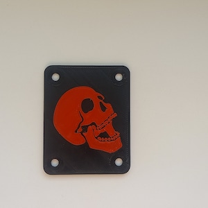 Op de afbeelding: Een zwart vierkant gitaarpedaal met een rode schedel afbeelding. De schedel is naar rechts gericht en heeft een zwarte omtrek.