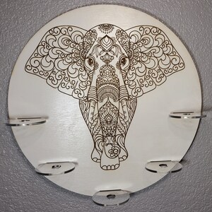 May include: A white circular wooden wall decor with a detailed mandala-style elephant design. The elephant is brown and has intricate patterns on its body and ears. There are five clear acrylic shelves around the edge of the circle.