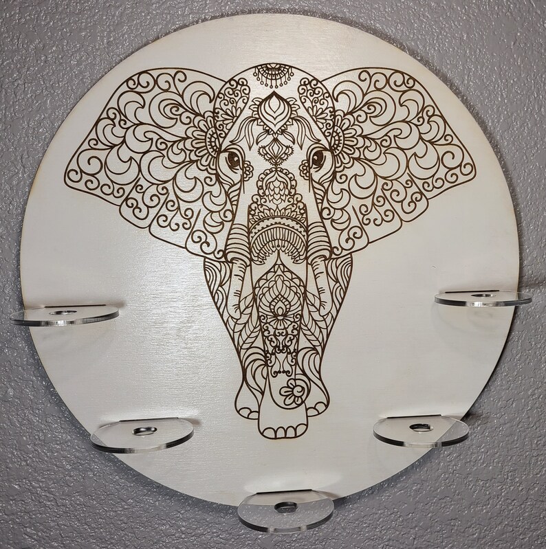 May include: A white circular wooden wall decor with a detailed mandala-style elephant design. The elephant is brown and has intricate patterns on its body and ears. There are five clear acrylic shelves around the edge of the circle.