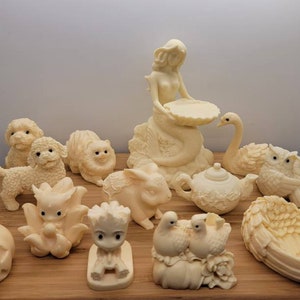 May include: A collection of 13 white ceramic figurines, including a mermaid, a swan, a teapot, a pumpkin, a rabbit, a cat, two owls, two dogs, a heart-shaped dish, and two birds. The figurines are arranged on a wooden surface.