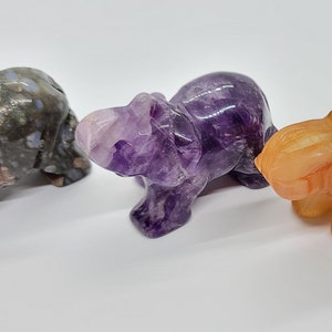 May include: Three carved elephant figurines made of different types of stone. The first elephant is made of a light gray and pink stone with brown and white markings. The second elephant is made of a purple amethyst stone. The third elephant is made of a light orange stone.