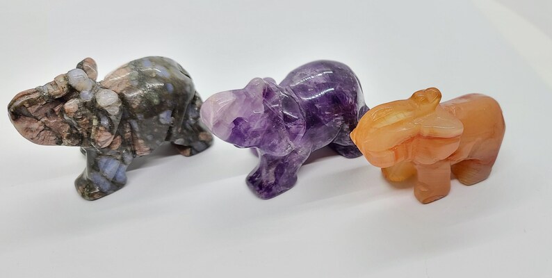 May include: Three carved elephant figurines made of different types of stone. The first elephant is made of a light gray and pink stone with brown and white markings. The second elephant is made of a purple amethyst stone. The third elephant is made of a light orange stone.