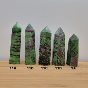 May include: Five green and pink gemstone points, labeled 11A, 11B, 11C, 11D, and 9A, are arranged in a row on a wooden surface.