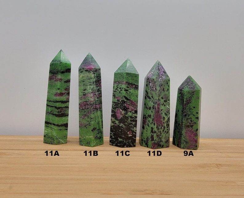 May include: Five green and pink gemstone points, labeled 11A, 11B, 11C, 11D, and 9A, are arranged in a row on a wooden surface.