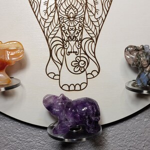 May include: A wooden mandala with a carved elephant design. There are five small elephant figurines made of different colored stones, including orange, purple, and gray, placed on the mandala.