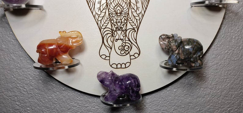 May include: A wooden mandala with a carved elephant design. There are five small elephant figurines made of different colored stones, including orange, purple, and gray, placed on the mandala.