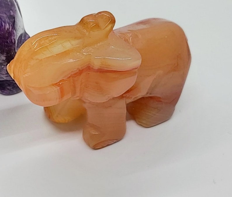 May include: A small, orange-colored, carved stone elephant figurine. The elephant has a smooth, polished surface and is standing on all four legs.