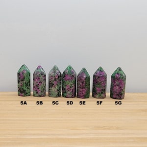 May include: Seven green and pink gemstone points, labeled 5A through 5G, arranged in a row on a wooden surface.