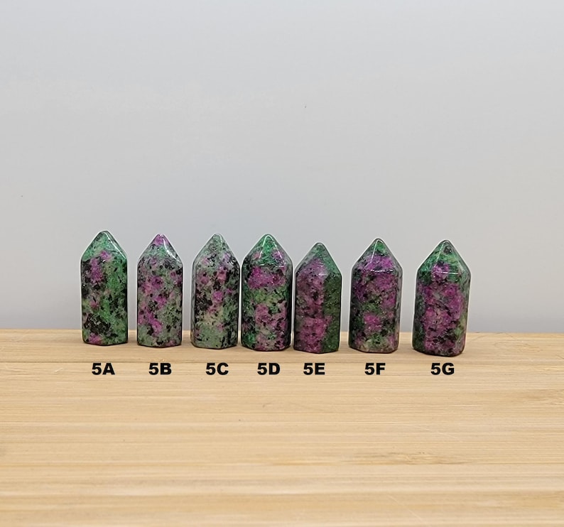 May include: Seven green and pink gemstone points, labeled 5A through 5G, arranged in a row on a wooden surface.