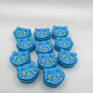 May include: A collection of ten small, light blue, cat-shaped containers. Each container features a printed face with yellow eyes, dark blue markings, and a white grin. The lids are shaped like cat ears, and the containers have a smooth, matte finish.