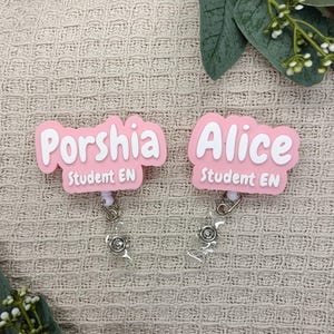 May include: Two custom badge reels featuring pink, cloud-shaped acrylic toppers with white lettering. The left badge reads Porshia Student EN, and the right badge reads Alice Student EN. Each includes a clear plastic strap and a metal snap button.