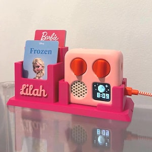 May include: A bright pink plastic organiser stand featuring the name Lilah in raised lettering, holding two cards labelled Barbie and Frozen. Next to it sits a light pink digital clock with two orange knobs, a speaker grille, and a digital display showing 8:09.