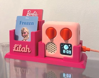 May include: A bright pink plastic organizer stand featuring the name Lilah in raised lettering, holding two cards labeled Barbie and Frozen. Next to it sits a light pink digital clock with two orange knobs, a speaker grille, and a digital display showing 8:09.