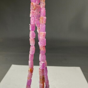 Può includere: Fili di perline di pietre preziose rosa e rosa-rossastro tagliate grezze. Le perline sono di forma quadrata e infilate insieme, tenute da una chiusura argentata. Le pietre preziose hanno un aspetto naturale, non lucido, con variazioni di colore e consistenza.
