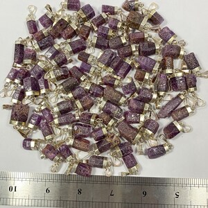 May include: A large group of purple and silver gemstone pendants. The pendants are approximately 1 inch long and are arranged in a pile. A ruler is visible in the bottom of the image, with markings from 6 to 10.