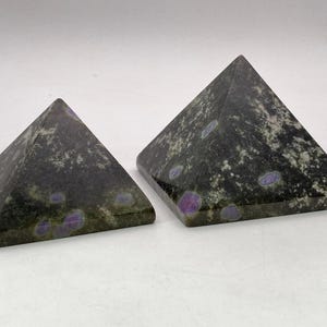 May include: Two dark green pyramid-shaped decorative objects with purple and white flecks. The pyramids have a smooth, polished surface and are displayed on a white background. The pyramids are likely made of stone or a similar material.