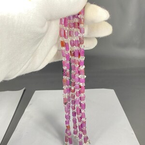 Può includere: Primo piano di più fili di perline di pietre preziose rosa e trasparenti. Le perline sono tenute da una mano guantata. Le perline rosa sono rettangolari e le perline trasparenti hanno una forma irregolare. I fili sono collegati in basso con chiusure argentate.