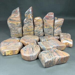May include: A collection of polished stones in various shapes and sizes. The stones display a mix of colors, including reddish-brown, yellow, and gray, with intricate patterns. Some stones are upright, while others are flat.
