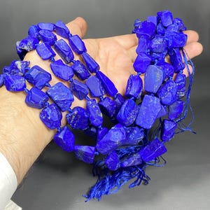 Könnte beinhalten: Eine Strähne tiefblauer Lapislazuli-Edelsteinkugeln, jede Kugel ist unregelmäßig geformt und hat eine raue, natürliche Textur. Die Kugeln sind auf einer blauen Schnur aufgefädelt und haben blaue Quasten an den Enden.