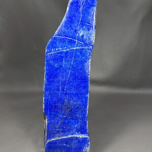 Puede incluir: Una losa alta y vertical de piedra lapislázuli que presenta un color azul profundo y vibrante con vetas de calcita blanca prominentes que corren horizontal y diagonalmente a través de la superficie pulida y lisa del espécimen mineral.