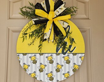 May include: A yellow and gray round door hanger with a black and yellow bow. The door hanger has a bee pattern and the text "Bee Happy".