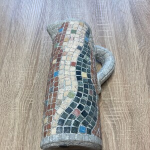 Könnte beinhalten: Eine Keramikkanne mit einem Mosaikdesign in verschiedenen Farben, darunter Rot, Grün, Blau und Weiß. Die Kanne hat einen Henkel und einen Ausguss.