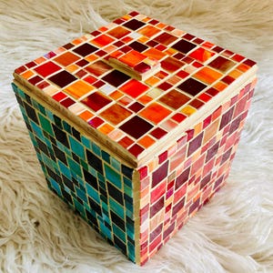 Könnte beinhalten: Eine quadratische Mosaikbox mit Holzrahmen und Deckel. Die Mosaikfliesen sind in Rot-, Orange- und Brauntönen auf der Oberseite und den Seiten gehalten, mit einem Abschnitt aus türkis- und grünen Fliesen auf einer Seite. Die Box steht auf einer weißen, flauschigen Oberfläche.