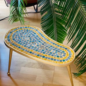 Könnte beinhalten: Kleiner, nierenförmiger Tisch mit einer Mosaikplatte in Blau- und Gelbtönen. Der Tisch hat ein goldfarbenes Metallgestell und Beine. Der Tisch ist ca. 61 cm lang, 30 cm breit und 46 cm hoch.