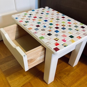 Könnte beinhalten: Weißer Beistelltisch mit einer Schublade und vier Beinen. Die Tischplatte ist mit einem Mosaik aus bunten, gemusterten Fliesen bedeckt.