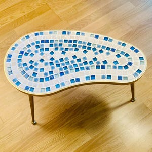 Könnte beinhalten: Ein Couchtisch im Retro-Stil mit einer Mosaik-Tischplatte. Der Tisch hat eine nierenförmige Form, goldfarbene Metallverkleidung und Beine. Die Mosaikfliesen sind weiß und in verschiedenen Blautönen. Ca. 100 cm lang.