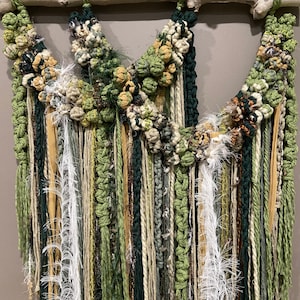 May include: A green, brown, and white macrame wall hanging with a wooden dowel at the top. The hanging features a variety of textures and materials, including yarn, string, and fabric.