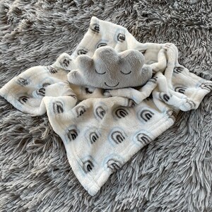 May include: A white baby blanket with a blue and gray rainbow pattern. A gray plush cloud with a smiling face is on top of the blanket.