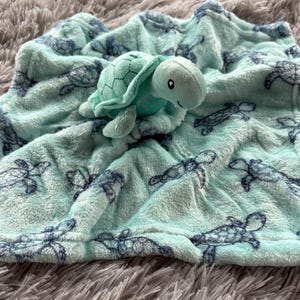 May include: A soft, turquoise baby blanket with a turtle design and a plush turtle toy. The blanket features a pattern of dark blue sea turtles and is made of a fuzzy material. The plush turtle is light turquoise.
