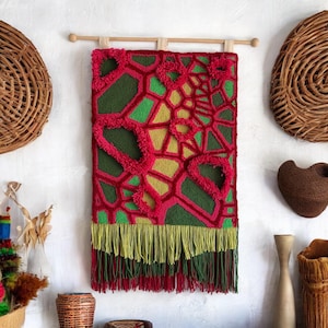 May include: A colorful wall tapestry with a geometric design in shades of pink, green, and yellow. The tapestry has a fringe border and is hanging on a wooden rod.