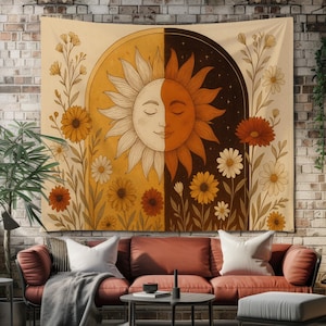 Pode incluir: Uma tapeçaria grande com um design dividido de um sol sorridente e uma noite estrelada. O sol é metade branco e metade laranja, com detalhes florais em tons de amarelo, laranja e castanho. A tapeçaria é exibida numa parede de tijolos.