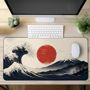 Puede incluir: Una alfombrilla de escritorio rectangular con una ilustración gráfica de una gran ola azul oscuro, un sol rojo prominente y un pico de montaña nevado. El diseño incluye pequeños pájaros negros sobre un fondo de color crema.