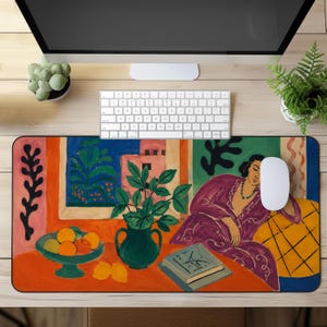 Puede incluir: Una alfombrilla de escritorio rectangular con una colorida ilustración pictórica de una persona con una túnica morada descansando junto a una mesa con un cuenco de naranjas, un jarrón verde con hojas verdes y un libro, sobre un fondo naranja y azul.