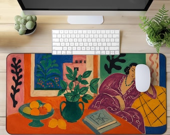 May include: A rectangular desk mat featuring a colorful, painterly illustration of a person in a purple robe resting near a table with a bowl of oranges, a green vase with green leaves, and a book, set against a vibrant orange and blue background.