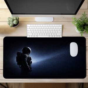 Può includere: Un tappetino da scrivania rettangolare con una grafica spaziale scura che raffigura un astronauta in tuta visto di profilo. Lo sfondo è nero intenso con un sottile effetto di luce blu e stelle sparse, offrendo un'ampia superficie per il mouse.