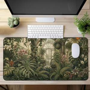 Könnte beinhalten: Eine rechteckige Schreibtischunterlage mit einer detaillierten botanischen Illustration eines Gewächshausinneren, gefüllt mit üppigen grünen Farnen, Monstera-Blättern, Hängepflanzen und verschiedenen blühenden Blumen in Rosa- und Weißtönen unter Bogenglasfenstern.