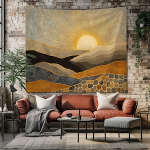 Op de afbeelding: Een groot wandtapijt met een gestileerd landschap met een opkomende zon. Het kunstwerk gebruikt tinten goud, bruin en grijs om bergen en een stralende zonnestralen af te beelden. Het wandtapijt wordt tentoongesteld aan een bakstenen muur boven een rode bank.