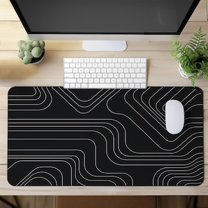 Può includere: Tappetino per mouse rettangolare nero con un design a linee bianche curve astratte. Un mouse e una tastiera bianchi sono sopra. Il tappetino è su una scrivania in legno chiaro, con un monitor di computer, un piccolo cactus e una felce sullo sfondo.