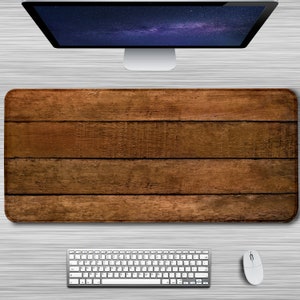 Puede incluir: Una alfombrilla de escritorio de madera marrón con un patrón de veta de madera. La alfombrilla está sobre un escritorio blanco con un monitor de ordenador plateado, un teclado blanco y un ratón blanco.