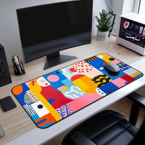 Op de afbeelding: Een kleurrijke abstracte geometrische design muismat met een blauw, rood, geel, roze en zwart kleurenschema. De muismat ligt op een bureau met een computermonitor en een computermuis.