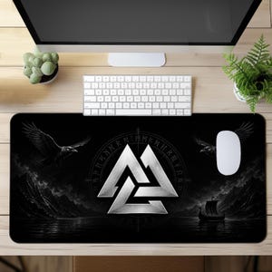 Pode incluir: Um tapete de secretária retangular com um design a preto e branco, apresentando um símbolo Valknut metálico central rodeado por um círculo rúnico. O fundo mostra uma paisagem escura com montanhas, um navio viking e dois corvos.