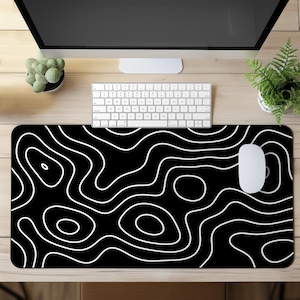 Könnte beinhalten: Schwarzes Mauspad mit weißem topografischem Design. Das Pad hat ein sich wiederholendes Muster aus weißen Konturlinien auf schwarzem Hintergrund. Eine weiße Computermaus und Tastatur liegen auf dem Holzschreibtisch.