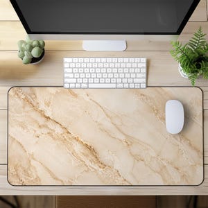 May include: A rectangular desk mat featuring a beige and brown marble pattern. A white computer mouse sits on the mat, alongside a white keyboard. A computer monitor, a small potted cactus, and a fern are also visible.