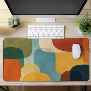 May include: A rectangular desk mat featuring an abstract pattern of overlapping rounded shapes in shades of olive green, teal, mustard yellow, burnt orange, and cream. The surface has a smooth texture and covers a large portion of the workspace.