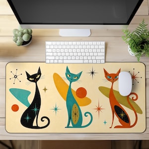 May include: A rectangular desk mat featuring a cream background with three stylised cats in black, teal, and orange. The design includes geometric shapes, starburst patterns, and circular accents in a mid-century inspired graphic illustration style.